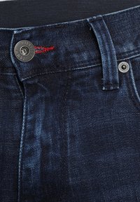 Jeans in denim blu scuro con bottone metallico inciso "Tommy Hilfiger", dettagli di cuciture rosse e design tradizionale a cinque tasche.
