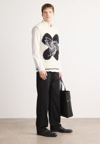 Pulover tricotat fără mâneci, de culoare crem, cu un design abstract negru, purtat peste o cămașă albă. Pantaloni negri și pantofi, ținând o geantă neagră.