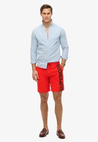 Camisa de botones azul claro, pantalones cortos de baño rojos con texto negro "SUPERDRY" y mocasines marrones. El modelo está de pie frente a un fondo neutro.
