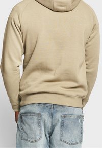 Felpa beige con cappuccio, di texture liscia e orlo a coste, indossata con jeans chiari strappati con tasche posteriori.