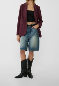 Bordeaux blazer over een zwarte crop top, gecombineerd met lichtblauwe denim bermuda shorts. Zwarte cowboylaarzen met reliëfdetails maken de look compleet.