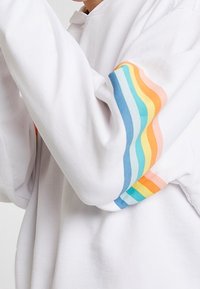Felpa bianca con dettagli a strisce multicolori in blu, arancione, giallo e verde sulle maniche. Tessuto morbido e vestibilità rilassata.