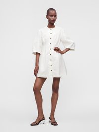 Mannequin mince portant une robe blanche boutonnée avec des manches aux coudes et des talons ouverts noirs, debout avec une main sur la hanche.