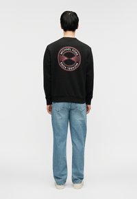 Μαύρο φούτερ με λογότυπο τυπωμένο στην πλάτη, συνδυασμένο με ανοιχτό μπλε τζιν παντελόνι και λευκά αθλητικά παπούτσια. Το φούτερ διαθέτει ριμπ μανσέτες και τελείωμα.