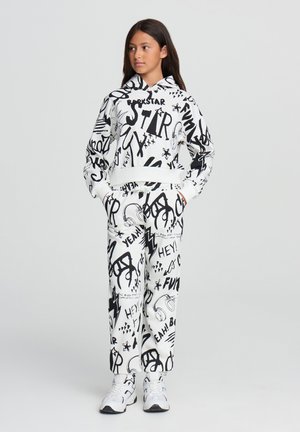 Completo con felpa bianca con cappuccio e pantaloni della tuta, decorato con disegni e testo in stile graffiti neri. Presenta un cappuccio e polsini elasticizzati. Le scarpe sono sneakers bianche.