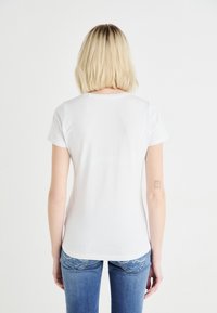 T-shirt blanc à manches courtes avec un col rond, design ajusté et texture lisse. Porté avec un jean en denim bleu, révélant un simple tatouage sur le bras.