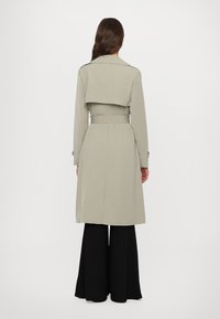 Trench-coat gris clair avec une ceinture, design croisé et détail d'un rabat à l'arrière, associé à un pantalon noir évasé.