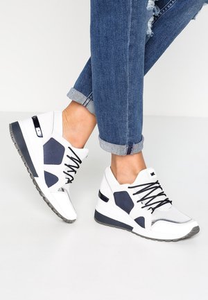 Personne portant des baskets blanches et bleu marine avec des lacets noirs et un jean bleu retroussé sur un fond blanc uni.