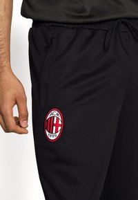 Svarta sportbyxor med ett nätmönster, utrustade med en röd och vit broderad AC Milan-logotyp på vänster lår. Dragsko i midjan ingår.