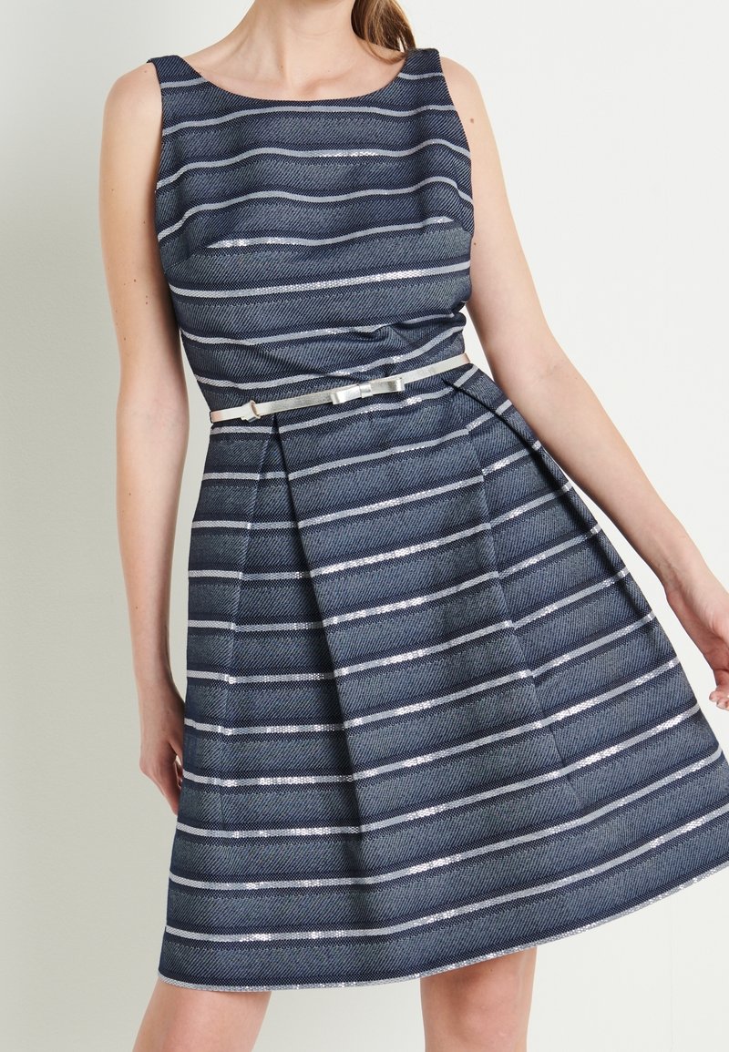 Femme portant une robe bleu marine sans manches avec des rayures argentées horizontales et une fine ceinture argentée avec un nœud à la taille.