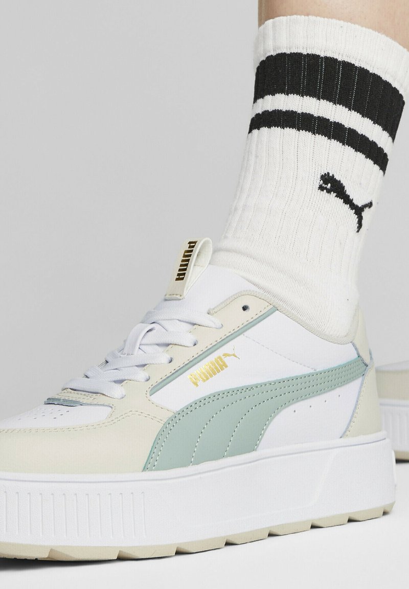 Bílé a světle mátové tenisky s robustní podrážkou, texturovanou kůží a zlatým logem. Nošeny s bílými ponožkami s žebrovaným vzorem a černými pruhy.