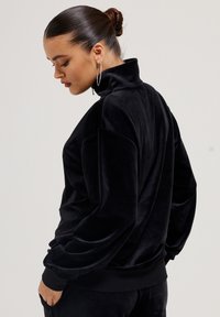 Vrouw met een hoge bun, die grote oorbellen en een zwarte fluwelen sweatshirt met hoge hals draagt, poseert met handen in haar zakken en kijkt naar beneden.