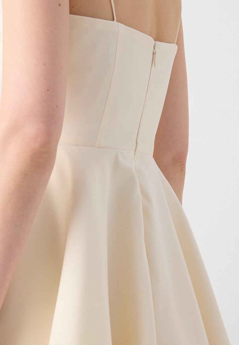 Helles beigefarbenes Kleid mit angepasstem Oberteil, dünnen Trägern und einem ausgestellten Rock. Verfügt über einen Reißverschluss an der Seite und einen glatten, strukturierten Stoff.