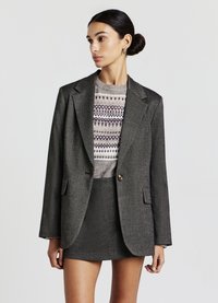 Blazer en laine grise avec des épaules structurées, fermeture à un bouton et poches avant. Porté sur un pull à motifs et une mini-jupe assortie.