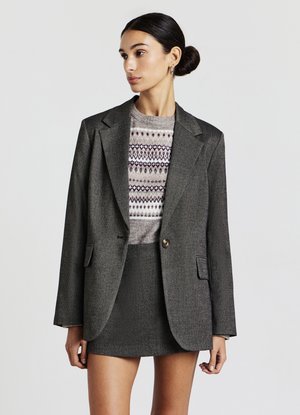 Blazer en laine grise avec des épaules structurées, fermeture à un bouton et poches avant. Porté sur un pull à motifs et une mini-jupe assortie.