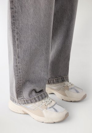 Jeans grigi a gamba larga indossati con scarpe da ginnastica atletiche in mesh bianche e beige su uno sfondo chiaro.