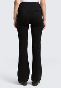 Schwarze hochgeschnittene Schlaghose mit zwei Gesäßtaschen, glatter Stofftextur und maßgeschneidertem Design. Gummi-sohlige Schuhe sind sichtbar.