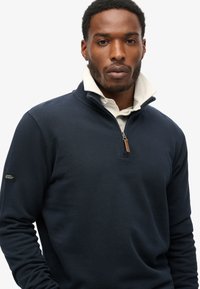 Sudadera navy con cuello de media cremallera, que presenta un cuello interior claro. Incluye un pequeño parche con el logo en la manga izquierda. Textura suave.
