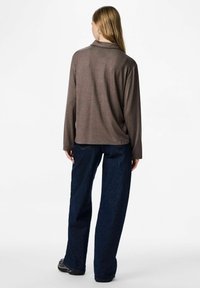 Žena okrenuta leđima, obučena u smeđu majicu dugih rukava, tamnoplave široke traperice i crne cipele na bijeloj pozadini.