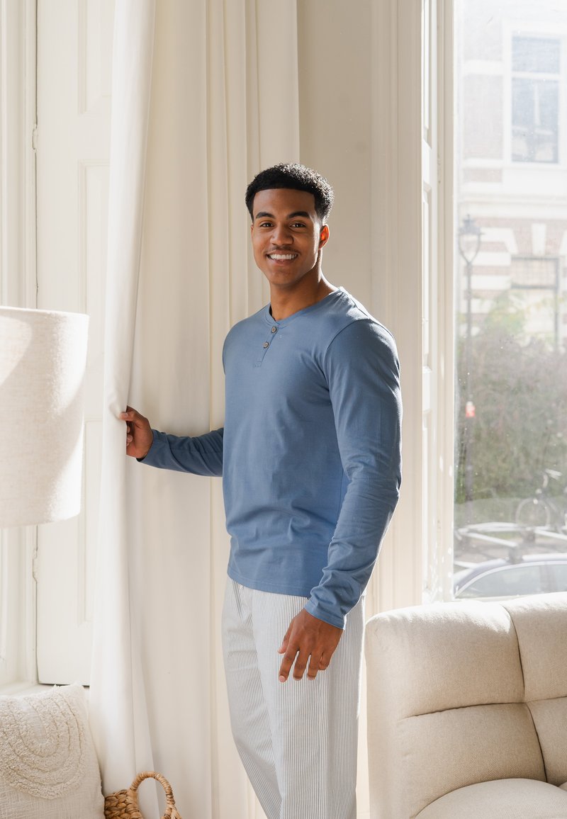Blauwe langemouwen henley shirt met drie knopen, gecombineerd met licht gestreepte broeken, in een heldere ruimte vlakbij witte gordijnen en een lamp.