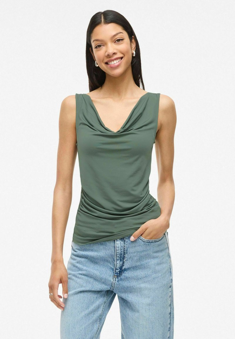 Top sans manches vert olive avec un col drapé et un détail froncé sur le côté, associé à un jean taille haute bleu clair. Texture du tissu lisse.
