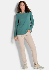 Eine Frau trägt ein türkisfarbenes Oberteil mit langen Ärmeln und Struktur, beigefarbene Slim-Fit-Hosen und beigetönige Sneaker. Sie steht mit einer Hand in der Tasche vor einem weißen Hintergrund.