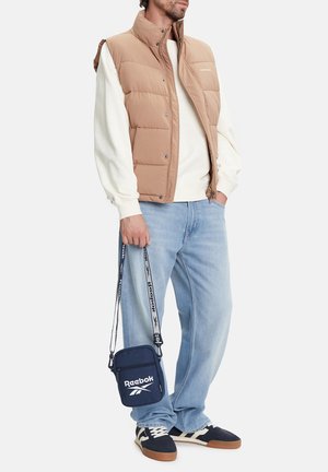 Mężczyzna w beżowej pikowanej kamizelce, białej bluzie, jasnoniebieskich dżinsach, czarnych trampkach, trzymający granatową torbę Reebok na ramię za pasek.