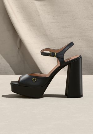 Mustad platvormi kõrge kontsaga sandaal pahkluu rihmaga, kuldse pandla ja väikese kuldse südamekujuga kaunistusega küljel, seistes beežil pinnal.