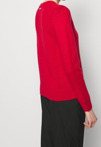 Czerwony sweter z dzianiny z okrągłym dekoltem, prążkowanymi mankietami i dołem. Widoczny detal z logo na plecach, wykonany z miękkiego, strukturalnego materiału.