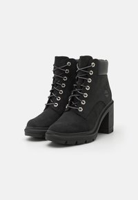 Black suede ankle boots with a chunky high heel, metal eyelets, and gray laces. Features a padded collar and lugged sole for traction.