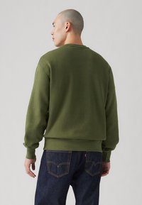 Olijfgroen sweatshirt met geribbelde manchetten en zoom, met een ronde hals en een ontspannen pasvorm. G gedragen met donkerblauwe jeans met achterzakken.