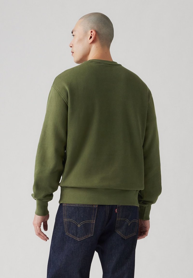 Sweatshirt vert olive à poignet et ourlet côtelés, avec un col rond et une coupe décontractée. Porté avec un jean bleu foncé affichant des poches arrière.