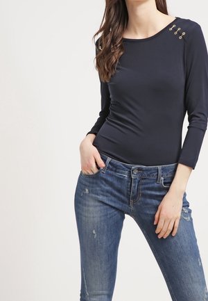 Woman wearing fitted navy blue long-sleeve top with gold button details on shoulder and distressed blue jeans, hands in front pockets.