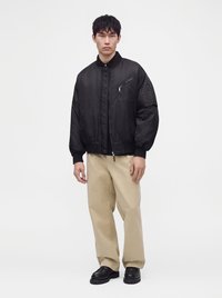 Νέος άνδρας που φοράει μαύρο bomber jacket, μπεζ παντελόνι με φαρδιά μπατζάκια και μαύρα παπούτσια στέκεται μπροστά σε λευκό φόντο.