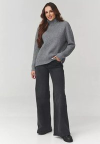 Kobieta z długimi brązowymi włosami, ubrana w szary sweter z golfem, czarne szerokie dżinsy i czarne buty, stojąca z jedną ręką w kieszeni.