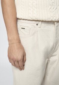 Cremfarbener Kabelstrickpullover, getragen über weißen, hoch taillierten, weit geschnittenen Hosen mit sichtbarer Fronttasche und silbernem Knopfdetail. Ein zartes Armband ziert das Handgelenk.