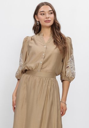 Vrouw draagt een beige jurk met knopen en opgepofte geborduurde mouwen, staat met ontspannen handen en kijkt lichtjes opzij.