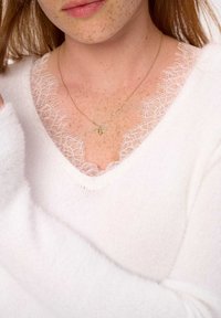 Witte trui met een V-hals, voorzien van een delicate kanten rand. Een gouden ketting met een bijpendant is zichtbaar tegen de huid.