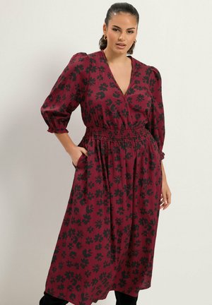 Femme portant une longue robe bordeaux à motif floral noir, manches bouffantes trois-quarts et taille élastique, debout avec une main dans la poche.