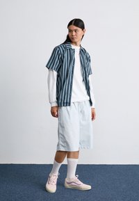 Jeune personne se tenant sur un tapis bleu, portant un short bleu clair, une chemise à manches longues blanche, une chemise à manches courtes rayée bleu, des chaussettes blanches et des baskets roses.