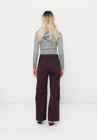 Kvinna med långt blont hår iklädd en grå huvtröja, mörk lila vida cargobyxor och svarta kitten heels, ståendes med ryggen vänd.