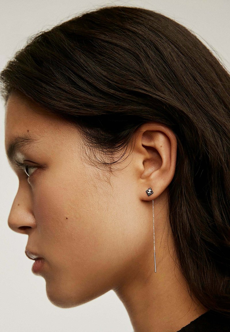 Pendientes de plata que presentan un stud cuadrado con una piedra transparente y una cadena de caída larga, delgada y recta. Cabello texturizado parcialmente visible.