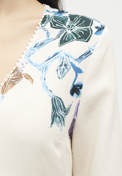 Gros plan d'un chemisier crème à col en V avec un motif floral bleu, vert, violet et marron sur la zone des épaules.