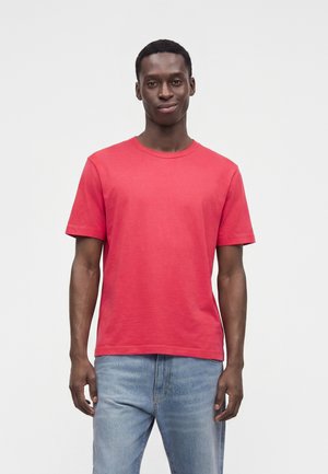 Homme portant un T-shirt rouge uni à manches courtes et un jean bleu clair, debout devant un fond blanc uni.