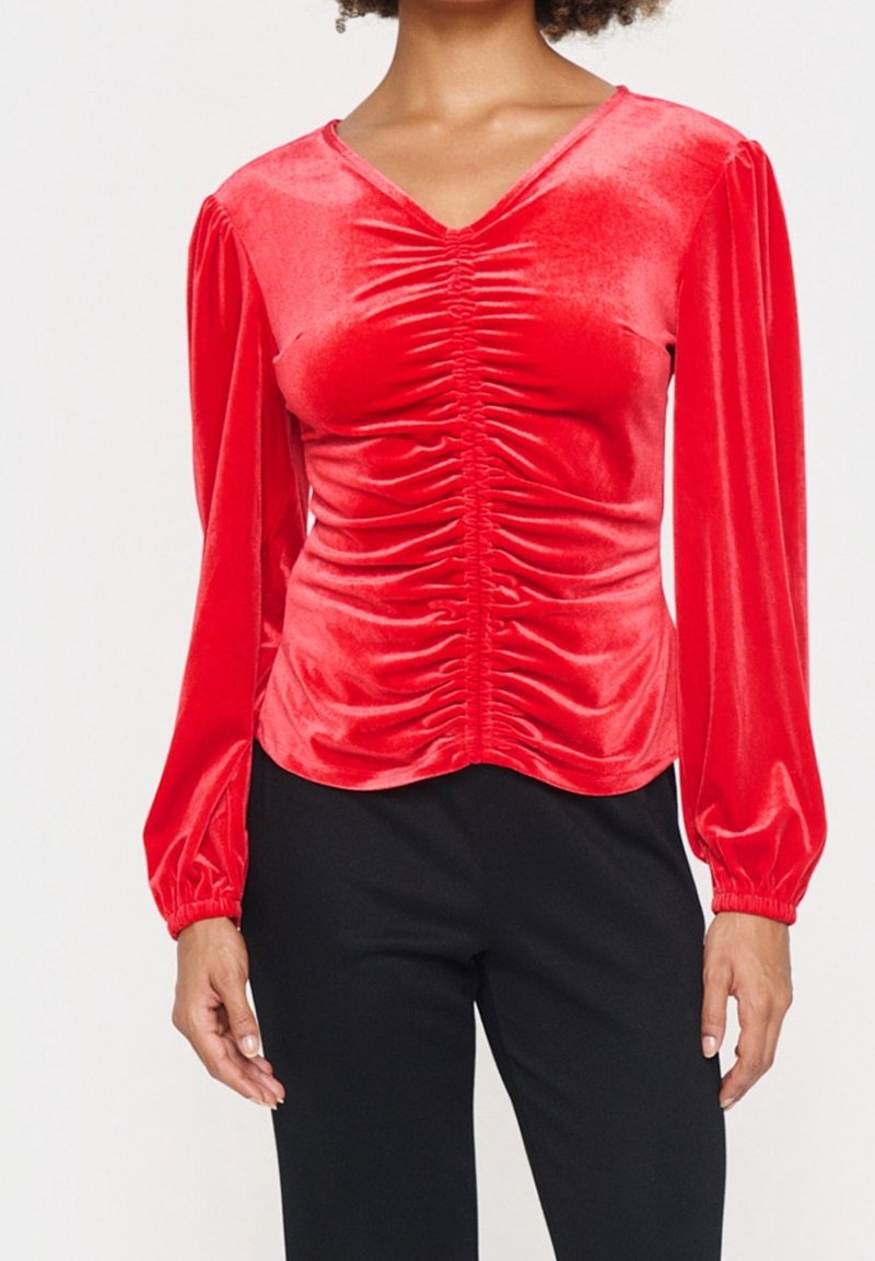 Haut en velours rouge avec de longues manches bouffantes, texture froncée à l'avant et décolleté en V. Silhouette ajustée, porté avec un pantalon sombre.