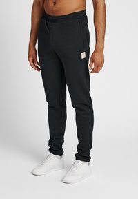 Schwarze Baumwoll-Sweatpants mit elastischem Bund, seitlichen Taschen und einem kleinen Etikett auf der Vorderseite. Kombiniert mit weißen Sneakers.