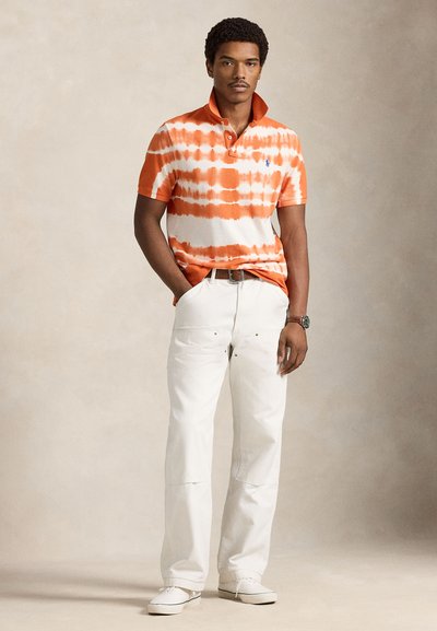 Πολύχρωμο πορτοκαλί και λευκό μπλουζάκι τύπου πόλο με γιακά, συνδυασμένο με παντελόνια σε εκρού χρώμα. Το μοντέλο φοράει λευκά παπούτσια. Απλό, καθημερινό ντύσιμο.