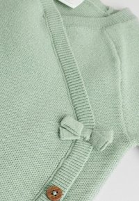 Zielony dzianinowy sweter z prążkowanym kołnierzem, z detalem w postaci kokardy i drewnianym guzikiem. Miękka tekstura i przytulny fason, idealny do noszenia w warstwach.