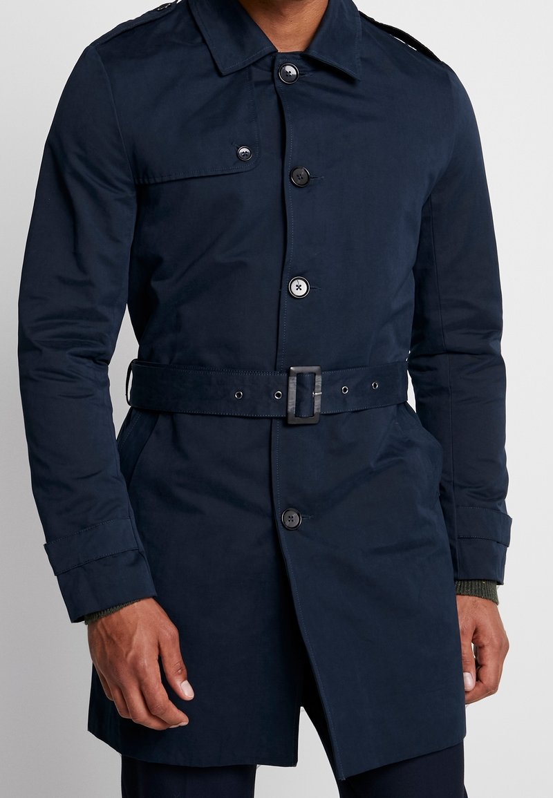 Homme portant un trench-coat bleu marine foncé ceinturé avec fermeture à boutons et poches latérales, debout devant un fond uni.