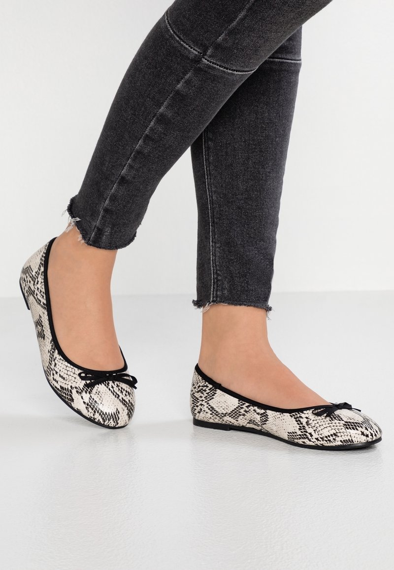 Personne portant un jean slim noir et des ballerines blanches à motif peau de serpent avec une bordure noire et de petits noeuds.
